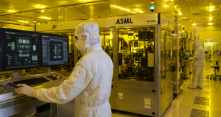 Cleanroom technicians wearing full protective bunny suits monitoring automation systems inside a modern semiconductor fabrication facility with yellow lithography lighting.
