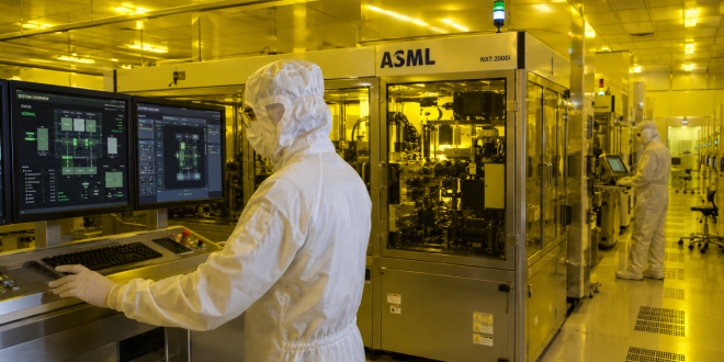 Cleanroom technicians wearing full protective bunny suits monitoring automation systems inside a modern semiconductor fabrication facility with yellow lithography lighting.