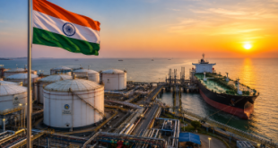 An Indian flag waving at a modern sea-facing petroleum terminal with huge white storage tanks, crude oil pipelines, and an ocean tanker docking in the background during a clear sunset.