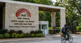 The stone brick entrance gate of the Indian Institute of Management Bangalore (IIMB) campus, featuring lush green trees and a cyclist passing by the main sign.