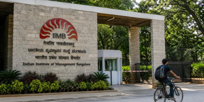 The stone brick entrance gate of the Indian Institute of Management Bangalore (IIMB) campus, featuring lush green trees and a cyclist passing by the main sign.