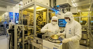 Clean-room engineers wearing protective suits handling a silicon semiconductor wafer inside an advanced manufacturing facility.