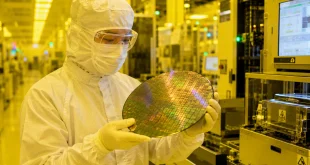 A semiconductor cleanroom technician wearing a full-body white sterile suit, protective mask, and goggles, carefully holding a reflective silicon microchip wafer under yellow cleanroom lighting inside a high-tech manufacturing fabrication facility.