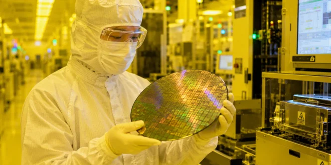 A semiconductor cleanroom technician wearing a full-body white sterile suit, protective mask, and goggles, carefully holding a reflective silicon microchip wafer under yellow cleanroom lighting inside a high-tech manufacturing fabrication facility.